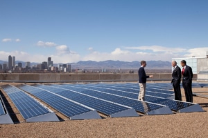 A group of people stand on a roof covered with solar panels with the Denver skyline in the background