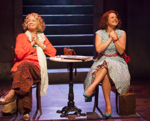 Two women sit with their legs crossed and hands clasped in excitement in Appoggiatura