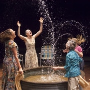 Two women and two children play in a small pool, splashing water high into the air in Benediction.
