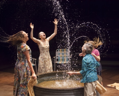 Two women and two children play in a small pool, splashing water high into the air in Benediction.