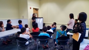 A group of students sit in chairs around a teacher at a white board