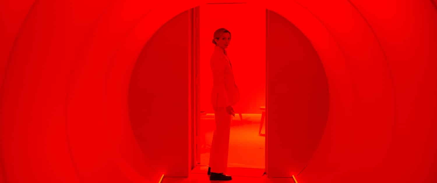 An individual in a white suit stands in front of a door at the end of a long hallway that is filled with red light