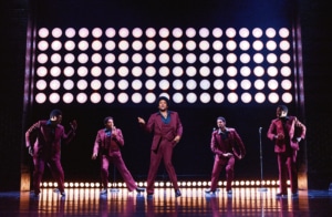 A group of five men in red suits sing and dance onstage in Ain't Too Proud