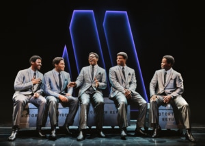 A group of men seated onstage sing together and smile in Ain't Too Proud