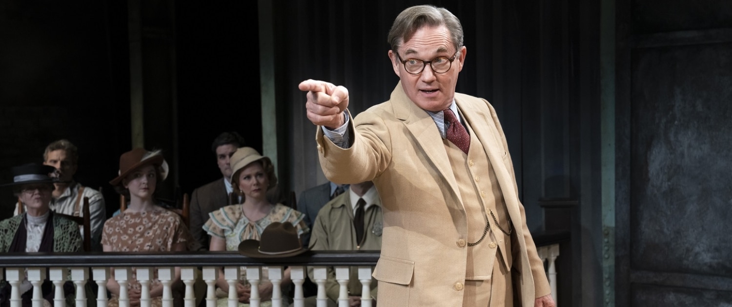 Richard Thomas, dressed in a tan three-piece suit, portrays Atticus Finch during a courtroom scene in To Kill a Mockingbird. The jury watches him from the background.