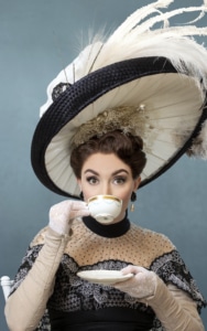 A woman sips from a cup of tea wearing a large feathered hat