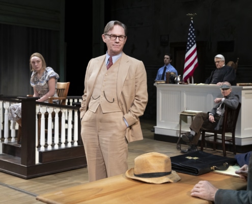 A woman in a floral dress sits at the witness stand before a judge, bailiff, and court reporter. Atticus Finch, centered with his hands in his pockets, looks towards the plaintiff.