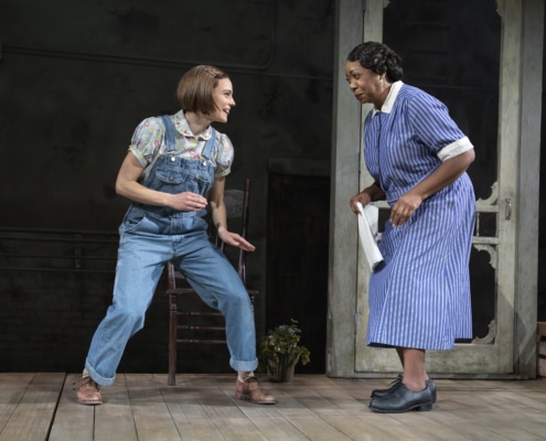 Melanie Moore (Scout Finch) appears at left in an athletic stance, grinning mischievously. Jacqueline Williams (Calpurnia) leans towards her and smiles broadly, dish towel in her right hand.