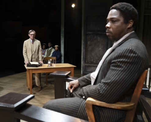 Richard Thomas (Atticus Finch) stands behind a desk in the courtroom as Yaegel T. Welch (Tom Robinson), wearing a black pinstripe on the witness stand, stares ominously to his left.