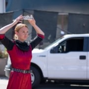 Female actor with blond hair in red costume dress holding a metal crown above her head with a serious expression on her face.