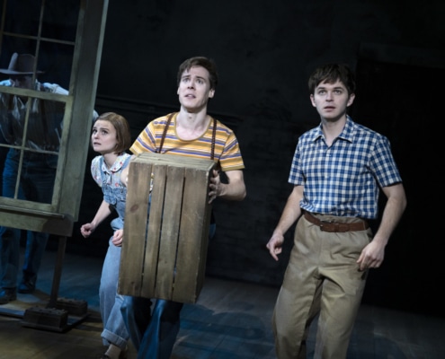 Travis Johns (Boo Radley) peers out from behind a dilapidated window as Melanie Moore (Scout Finch), Steven Lee Johnson (Dill Harris), and Justin Mark (Jem Finch) stand on his porch frozen in fear.