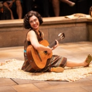 A woman sits onstage with a guitar in Much Ado About Nothing