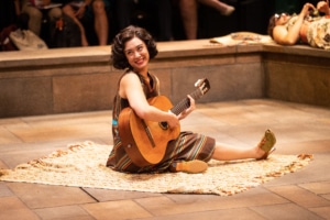 A woman sits onstage with a guitar in Much Ado About Nothing