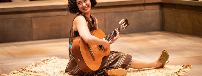 A woman sits onstage with a guitar in Much Ado About Nothing