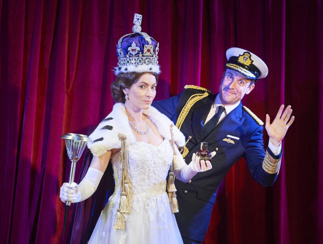 A woman dressed as Queen Elizabeth stands while a man waves from behind her