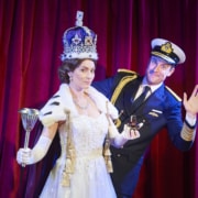 A woman dressed as Queen Elizabeth stands while a man waves from behind her