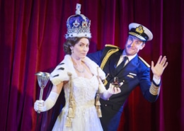 A woman dressed as Queen Elizabeth stands while a man waves from behind her