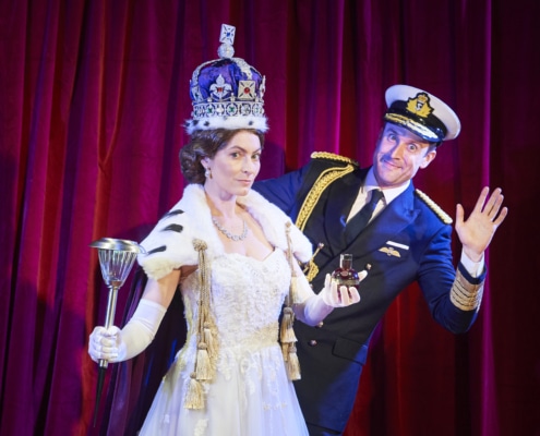 A woman dressed as Queen Elizabeth stands while a man waves from behind her