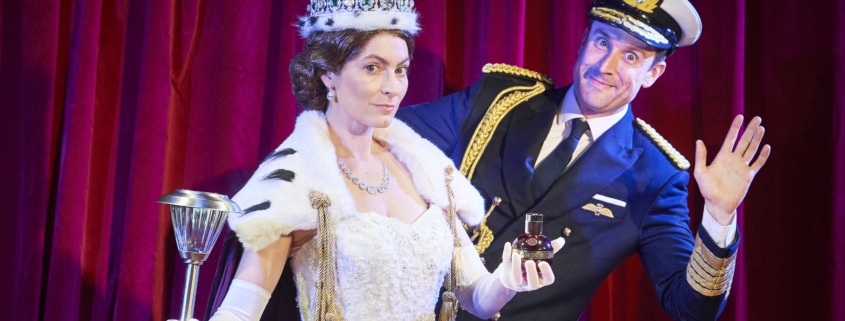 A woman dressed as Queen Elizabeth stands while a man waves from behind her