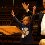 A young girl stands next to a conductor, both with the arms in the air