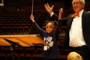 A young girl stands next to a conductor, both with the arms in the air