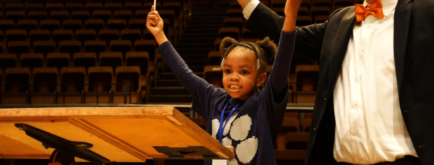 A young girl stands next to a conductor, both with the arms in the air