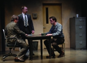 Two men sit while one stands at a table in The Pillowman