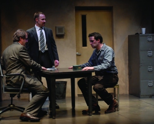 Two men sit while one stands at a table in The Pillowman