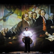 A woman stands onstage in front of Revolutionary War artwork in 1776