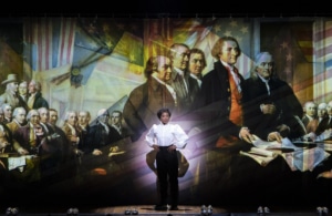 A woman stands onstage in front of Revolutionary War artwork in 1776