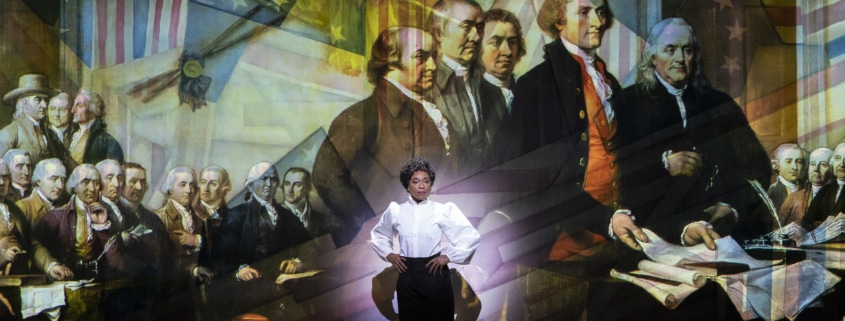 A woman stands onstage in front of Revolutionary War artwork in 1776