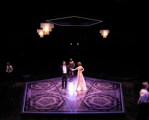 Three people in formal dress dance around the floor. At the center of the floor a woman in a pink floor length gown and a man in a tuxedo dance.