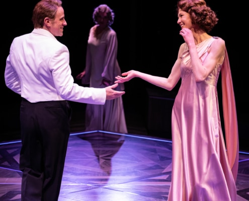 A woman in a pink floor length gown smiles and dances with a man in a white tuxedo coat.