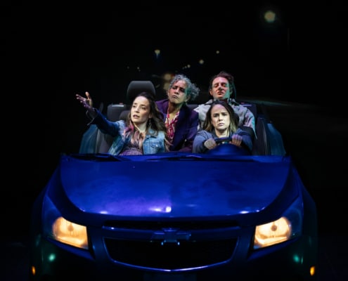 Three sisters ride in a car together. Celia drives as Yolie gestures at something out the window from the front passenger seat. Tere leans forward to see from her seat in the back next to the dead body.