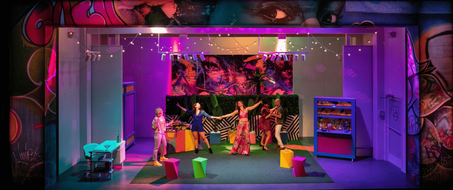 The cast of Laughs in Spanish dance on a set designed to look like a vibrant courtyard behind an art gallery. A bar with bottles of liquor and a table with an elaborate spread of food appear behind them.