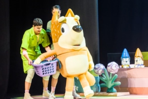 Mum is carrying a laundry basket, supported by actors in green jumpsuits.