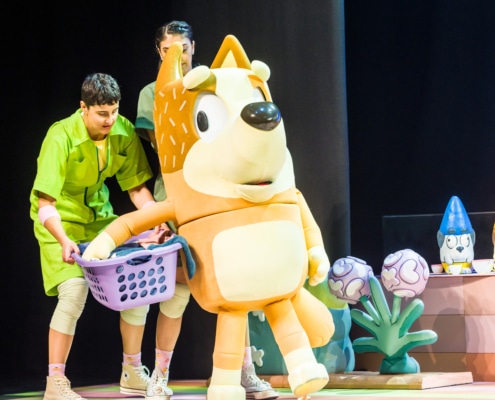 Mum is carrying a laundry basket, supported by actors in green jumpsuits.
