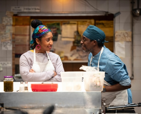 Two people chat in a kitchen. They are both wearing bandanas and aprons.