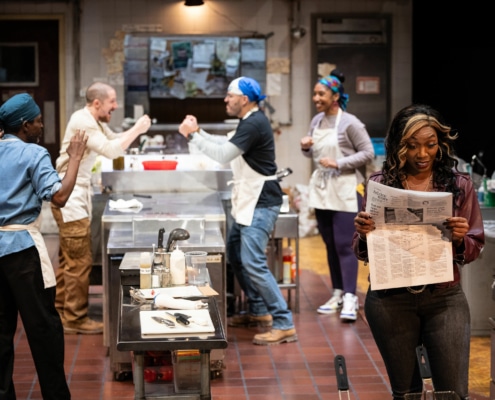 Four cooks, wearing aprons and bandanas, cheer in the background of the kitchen while a woman reads a newspaper in the foreground.