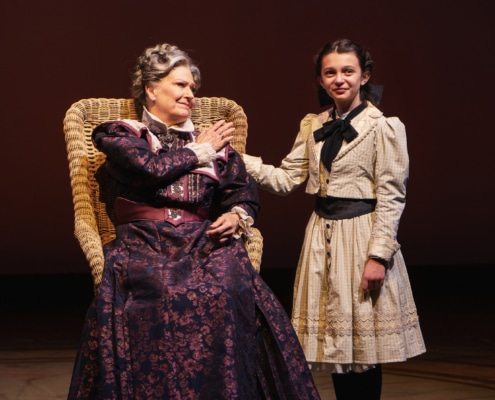 Sydney D’Angelo stands next to Leslie Alexander, sitting in a wicker wheelchair, with her hand on her shoulder.