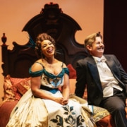 Soara-Joy Ross and Edward Staudenmayer laugh on an intricate bed in elegant costumes.