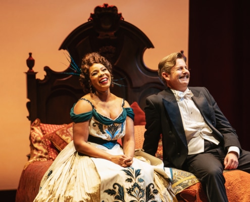 Soara-Joy Ross and Edward Staudenmayer laugh on an intricate bed in elegant costumes.