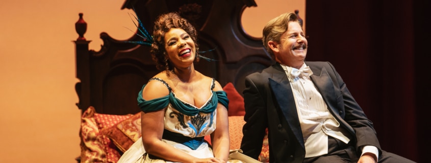 Soara-Joy Ross and Edward Staudenmayer laugh on an intricate bed in elegant costumes.