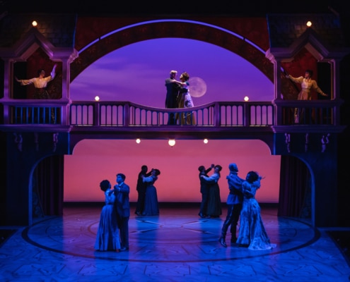 The cast of A Little Night Music waltz on a stage in elegant costumes as the moon sits behind them in a purple and pink sky.