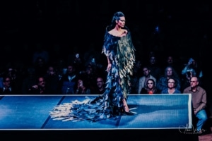 A model stands at the end of a catwalk wearing a blue, feathered style gown