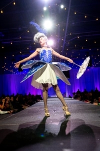 A model poses on the catwalk wearing a short, corseted dress. She holds a parasol out with extended arms.