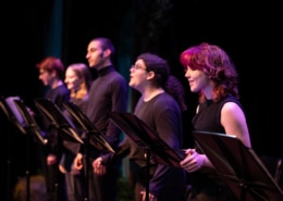 A row of actors standing at music stands wearing all black