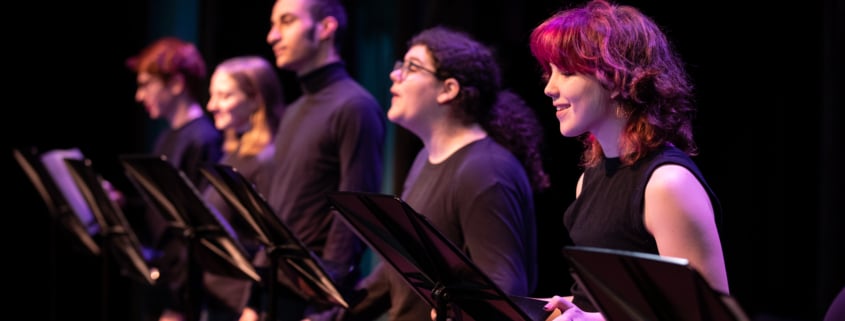 A row of actors standing at music stands wearing all black