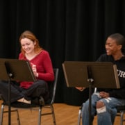 Two actors seated at music stands laugh together during rehearsal