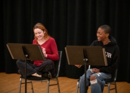 Two actors seated at music stands laugh together during rehearsal